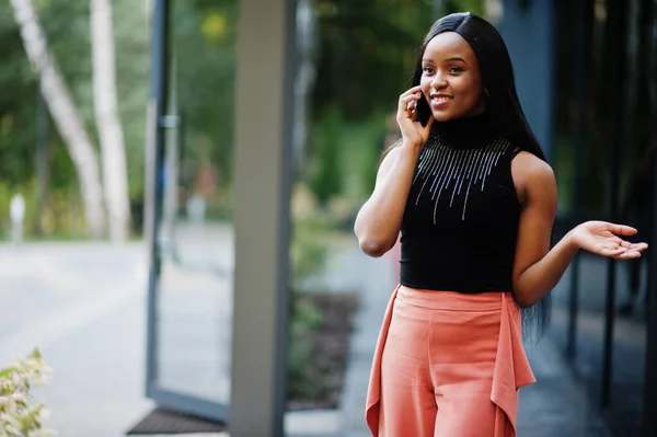 Fashionable african american woman in peach pants and black blouse speak on phone outdoor.