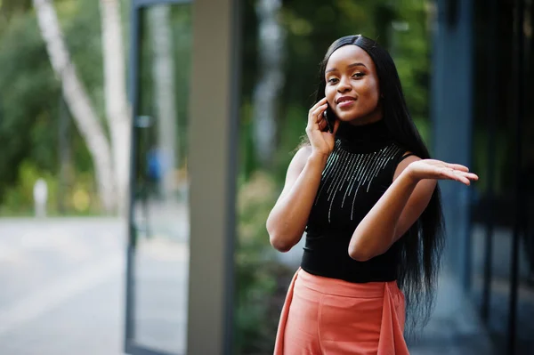 Fashionable african american woman in peach pants and black blouse speak on phone outdoor.