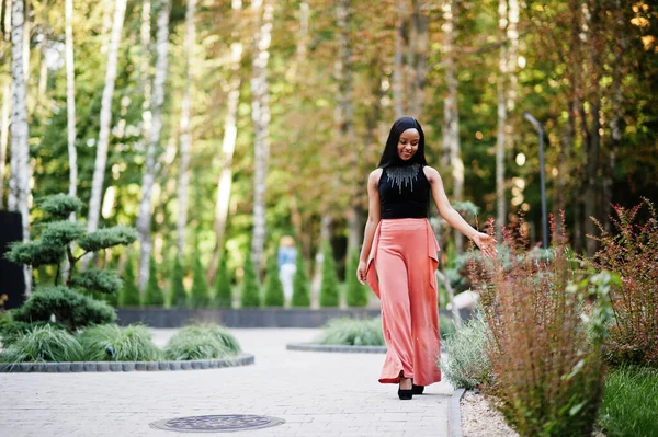 Fashionable african american woman in peach pants and black blouse pose outdoor.