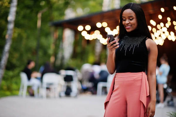 Fashionable african american woman in peach pants and black blouse look at mobile phone.