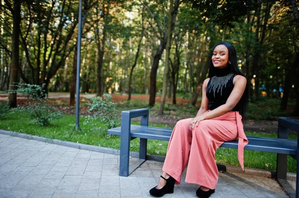 Fashionable african american woman in peach pants and black blouse pose outdoor, sitting on bench.