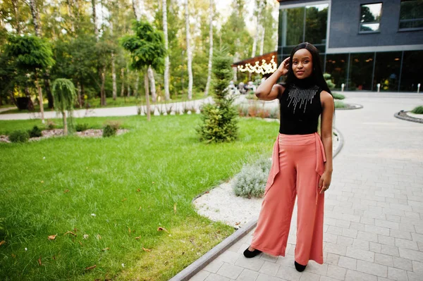 Fashionable african american woman in peach pants and black blouse pose outdoor.