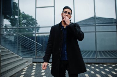 Orta Doğulu girişimci siyah ceket ve mavi gömlek giyer, ofis binasında sigara içerken gözlük takar..