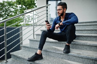 Orta Doğulu girişimci mavi gömlek giyer, ofis binasında gözlük takar ve cep telefonuna bakar..