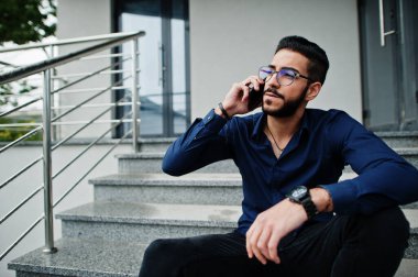 Orta Doğulu girişimci mavi gömlek giyer, ofis binasına karşı gözlük takar merdivenlerde oturur ve telefonla konuşur..