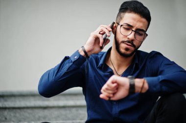 Orta Doğulu girişimci mavi gömlek giyer, ofis binasına karşı gözlük takar merdivenlerde oturur ve telefonla konuşur..