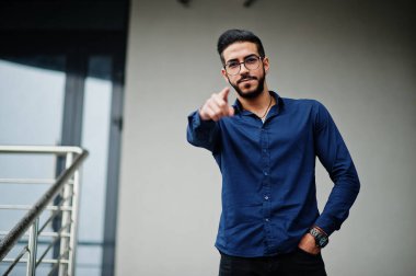 Orta Doğulu girişimci mavi gömlek giyer, ofis binasına karşı gözlük takar ve kameraya parmağını gösterir..