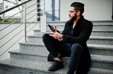 Orta Doğulu girişimci siyah ceket ve mavi gömlek giyiyor, ofis binasına karşı dizüstü bilgisayarlı gözlük takıyor.