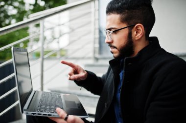Orta Doğulu girişimci siyah ceket ve mavi gömlek giyiyor, ofis binasına karşı dizüstü bilgisayarlı gözlük takıyor.