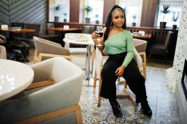 Yeşil kazak ve siyah kot giyen çekici Afro-Amerikan kadının portresi restoranda elinde bir bardak kırmızı şarapla poz veriyor..