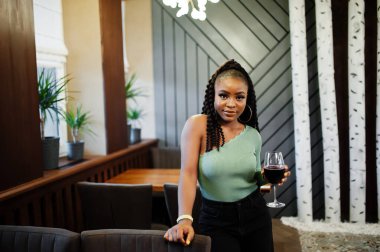 Yeşil kazak ve siyah kot giyen çekici Afro-Amerikan kadının portresi restoranda elinde bir bardak kırmızı şarapla poz veriyor..
