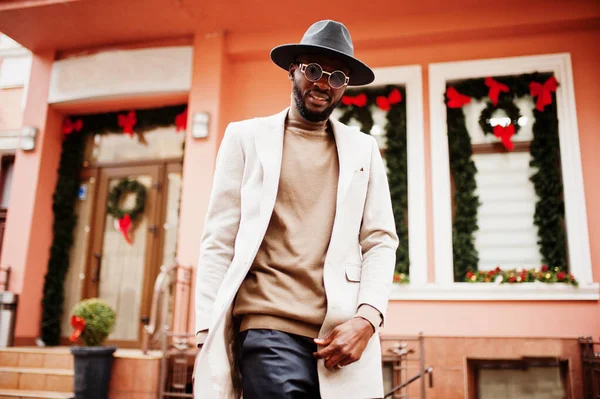 スタイリッシュなアフリカ系アメリカ人男性は新しい年の装飾と花輪と家に対してサングラスポーズとベージュのジャケットと黒の帽子を着用 — ストック写真