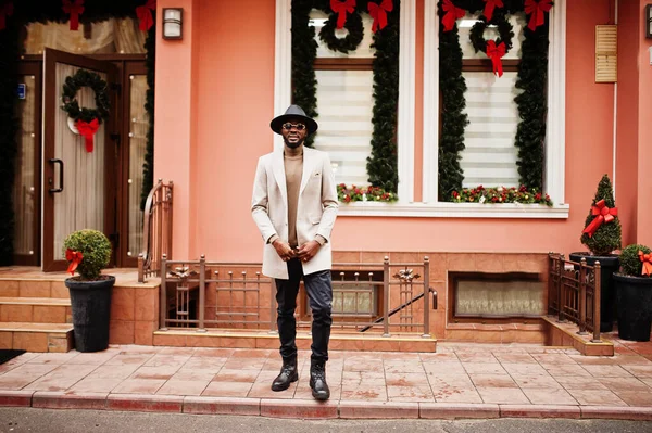 スタイリッシュなアフリカ系アメリカ人男性は新しい年の装飾と花輪と家に対してサングラスポーズとベージュのジャケットと黒の帽子を着用 — ストック写真