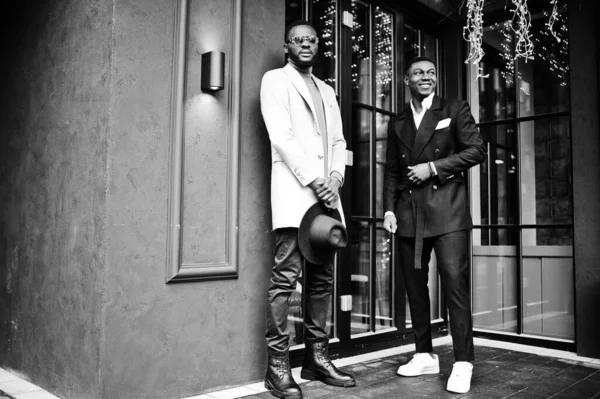 Two fashion black men pose against house with garlands. Fashionable portrait of african american male models. Wear suit, coat and hat.