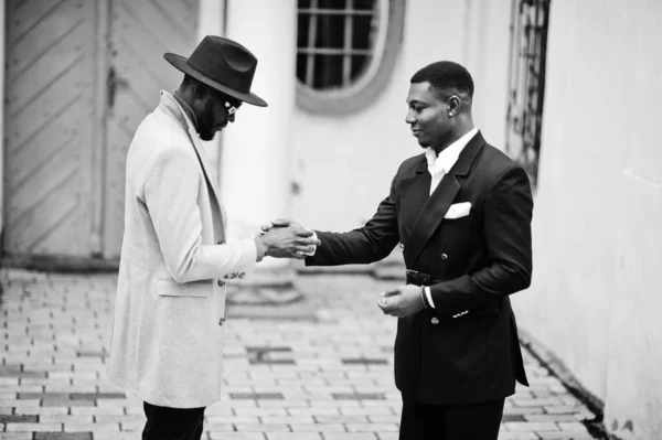 Two fashion black men shake hands each other. Fashionable portrait of african american male models. Wear suit, coat and hat.