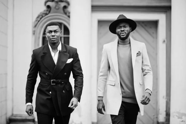 Two fashion black men walking on street. Fashionable portrait of african american male models. Wear suit, coat and hat.