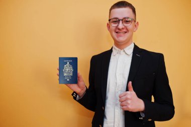 Sarı arka planda Kanada pasaportu tutan yakışıklı genç adam, mutlu ve başparmağını kaldır. Amerika ülkelerine seyahat.