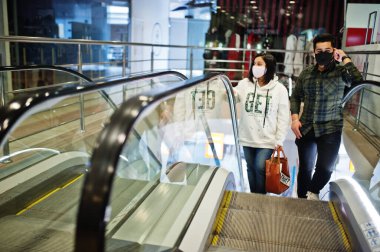 Asyalı çift maske takıyor. Ticaret merkezinde yürüyen merdivene tırmanıyorlar..