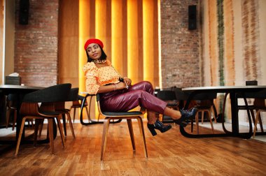 Kırmızı Fransız bereli zarif Afro-Amerikan kadın, büyük altın zincirli polka bluz ve deri pantolon, iç mekânda poz veriyor..