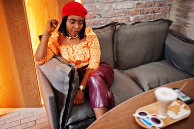 Kırmızı Fransız bereli zarif Afro-Amerikan kadın, büyük altın boyunlu polka bluz ve deri pantolon restoranda oturuyor ve latte içiyor..