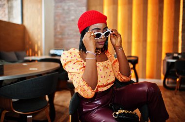 Kırmızı Fransız bereli zarif Afro-Amerikan kadın, büyük altın boyunlu zincir, güneş gözlüğü, benekli bluz ve deri pantolon..