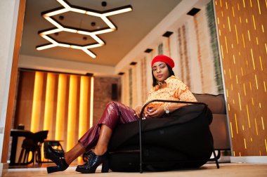 Kırmızı Fransız bereli zarif Afro-Amerikan kadın, büyük altın zincirli polka bluz ve deri pantolon pouf 'un üzerinde oturuyor..