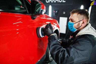 Araba temizleme konsepti. Yörünge parlatıcısı olan maskeli bir adam tamircide turuncu SUV araba cilalıyor..