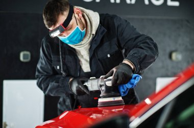 Araba temizleme konsepti. Yörünge parlatıcısı olan maskeli bir adam tamircide turuncu SUV araba cilalıyor..