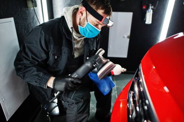 Araba temizleme konsepti. Yörünge parlatıcısı olan maskeli bir adam tamircide turuncu SUV araba cilalıyor..