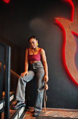 Kırmızı Marsala bluzlu ve kotlu Afrikalı kadın duvarda alevlerle poz vermiş..