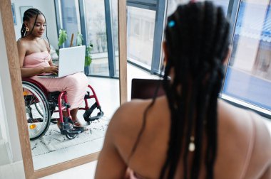 Genç, engelli, tekerlekli sandalyede oturan, aynaya karşı dizüstü bilgisayarla çalışan Afro-Amerikan kadın..