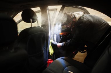 Üniformalı ve solunum cihazlı bir adam. Araba yıkama merkezinde çalışıyor. İçini sıcak buhar temizleyicisiyle temizliyor. Araba temizleme kavramı.