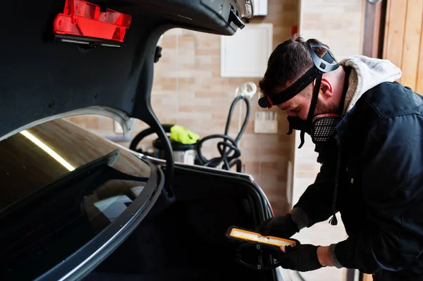 Man in uniform and respirator, worker of car wash center, cleaning car interior with hot steam cleaner. Car detailing concept.