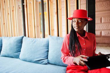 Pretty braids business african american lady bright bossy person friendly wear office red shirt, hat and trousers, hold tablet on hands.