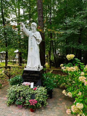 Banneux, Belçika - 20 Ekim 2023: Beyaz mermer Aziz Charbel heykeli fener tutan bir keşiş çiçek açan bitkilerle dolu bir bahçede duruyor. Banneux Leydisinin Sığınağı