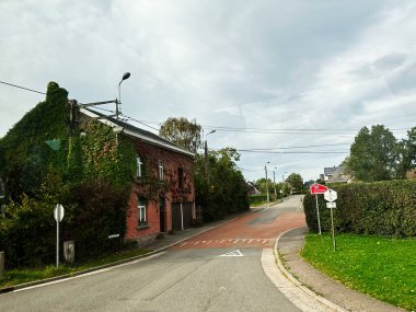 Banneux, Belçika - 20 Ekim 2023: Sarmaşık kaplı tuğla ev, çitler ve trafik işaretlerinin yer aldığı sakin bir Belçika sokak sahnesi.