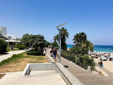 Paralimni, Kıbrıs - 18 Temmuz 2024: Kıbrıs Rum Kesimi 'nin canlı Protaras Sahili, mavi su ve berrak gökyüzünün yanında ziyaretçiler geziniyor.