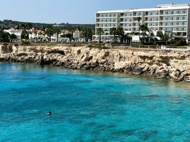 Paralimni, Kıbrıs - 18 Temmuz 2024: Kıbrıs Rum Kesimi 'nin Paralimni kentinde günışığı kıyı sahnesi modern bir Sunrise Jade oteli, berrak mavi su ve kayalık kıyı manzarası.
