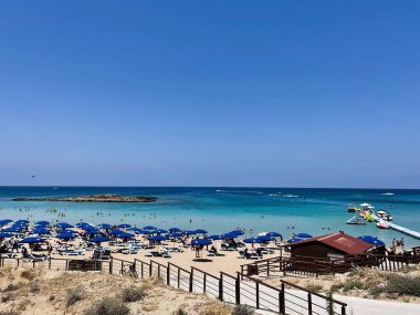 Paralimni, Kıbrıs - 18 Temmuz 2024: Kıbrıs Rum Kesimi 'nin Paralimni kentindeki güneşli sahneler, mavi sular, şemsiyeler ve canlı bir kıyı şeridinin keyfini çıkaran insanlar.