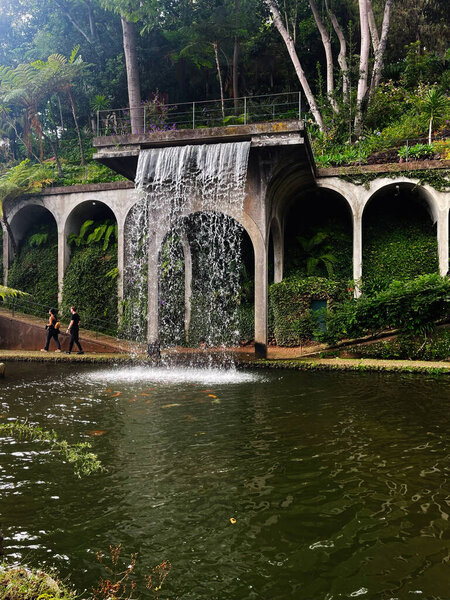 Water cascades from a concrete terrace into a pond surrounded by arches, plants, and visitors.