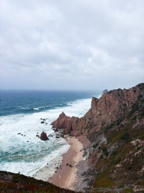 Engebeli kızıl kayalıklar tenha bir sahil olan Roca Burnu ile karamsar gökyüzünün altında dalgalar çarpışıyor..