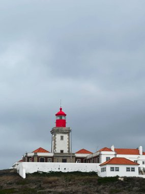 Coastal Cabo da Roca Deniz feneri sahnesinde parlak kırmızı bir deniz feneri ve deniz kenarında turuncu çatıları olan beyaz duvarlı binalar var..