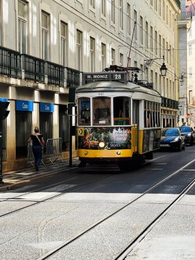 Lizbon, Portekiz - 28 Eylül 2025: Klasik bir sarı Lizbon Tramvayı güneş ışığı altındaki mağaza önleri ve yayaların yanında seyahat ediyor.