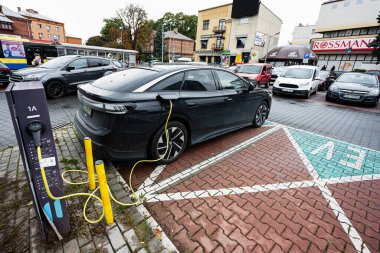 Tarnow, Polonya - 10 Ekim 2025: Pürüzsüz bir Volkswagen ID.7 sedan, şehir dükkanları ve park halindeki arabaların ortasındaki şarj direğine bağlanıyor.