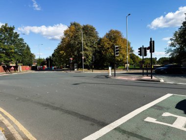 Sonbahar ağaçlarının altında trafik ışıkları, arabalar ve yayaların yer aldığı hareketli bir Manchester sokak sahnesi.