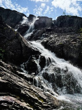 Güçlü bir şelale parlak bir gökyüzünün altındaki karanlık kayaların üzerinden akar, hareketi ve doğal dramı yakalar..