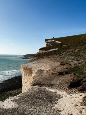 Seven Sisters Kayalıkları 'ndaki sahil uçurumu sahnesi mavi denizi, çakıl taşını ve uzak bir yürüyüşçüyü gösteriyor..