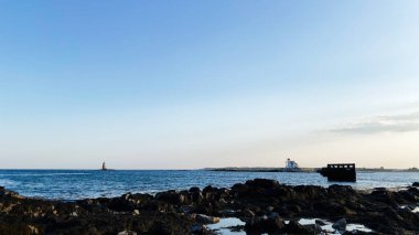 Balina Sırtı Deniz fenerindeki sahil manzarası. Kayalar, sakin su ve sahil kenarında uzak bir deniz feneri..