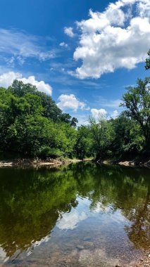Kinkaid Gölü 'nün sakin akıntısı yeşil ormanın içinden bükülür parlak bulutların altında, gökyüzünü ve çevresini çevreleyen yaprakları yansıtır.