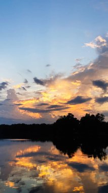 Altın gün batımı bulutları sakin bir göl üzerinde boyar, sıcak renkleri ve kıyı boyunca siluetli ağaçları yansıtır..